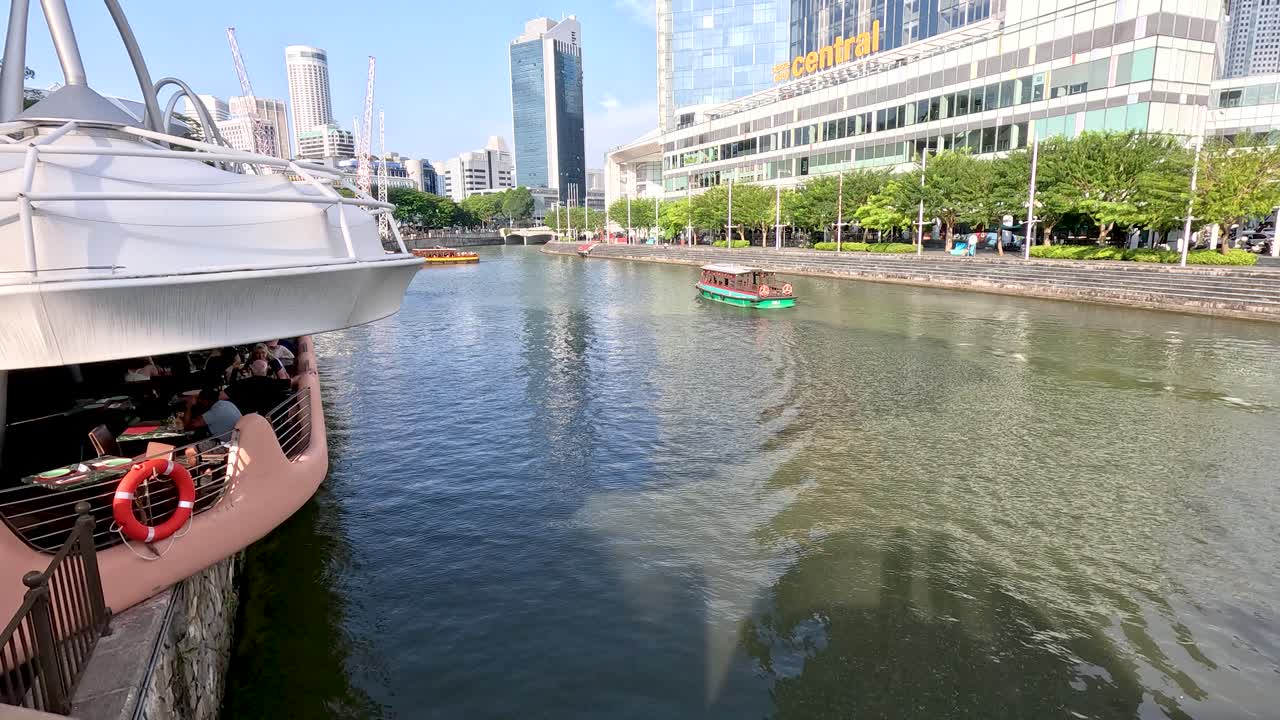 Camera pans along river, passing boats, waterfront buildings, and high-rise architecture in bright daylight