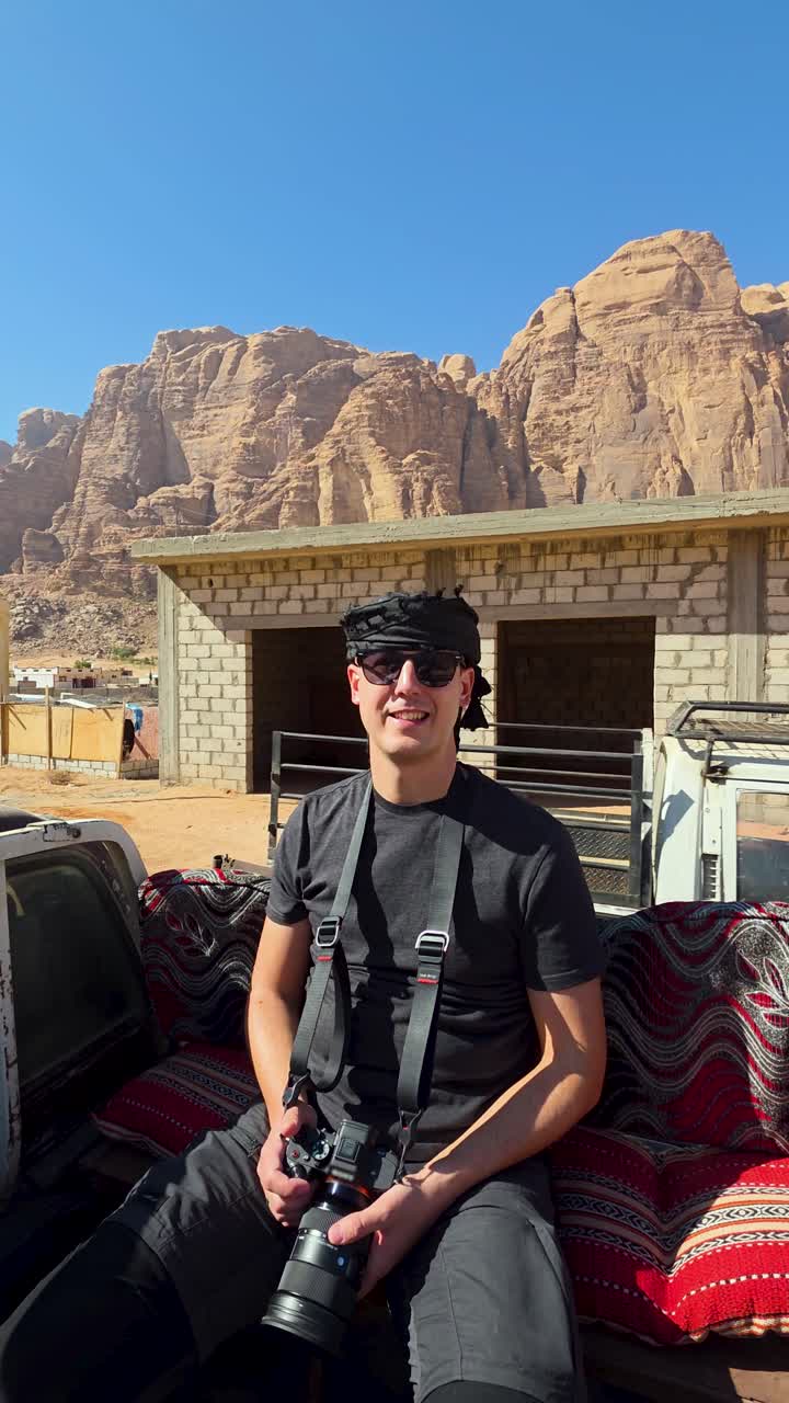Um fotógrafo masculino usando um lenço de cabeça tradicional sentado na traseira de uma picape durante um safári no deserto em Wadi Rum, Jordânia. Ideal para vlogs de viagem e documentários de aventura