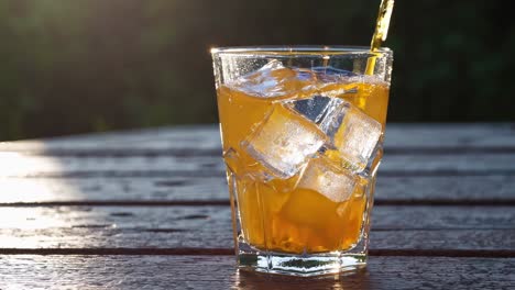 Orange juice is being poured into a glass filled with ice cubes, placed on a wooden table, creating a refreshing summer beverage under the sunlight