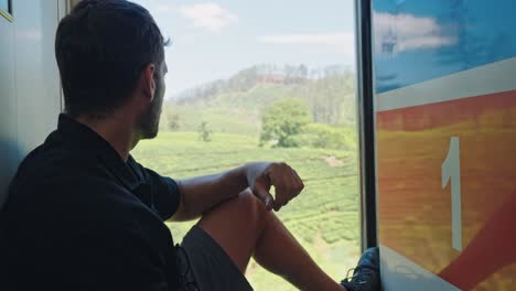 a man sits casually on the open door of a train traveling the scenic ella to kandy route in sri lanka.