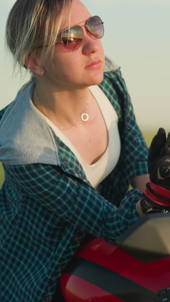 한 젊은 여성이 오토바이 거울에서 그녀의 반사를 보면서 선글라스를 조정합니다. 그녀는 개방된 필드의 부드러운 배경과 자전거의 은 몸으로 사려 깊은 것처럼 보입니다.