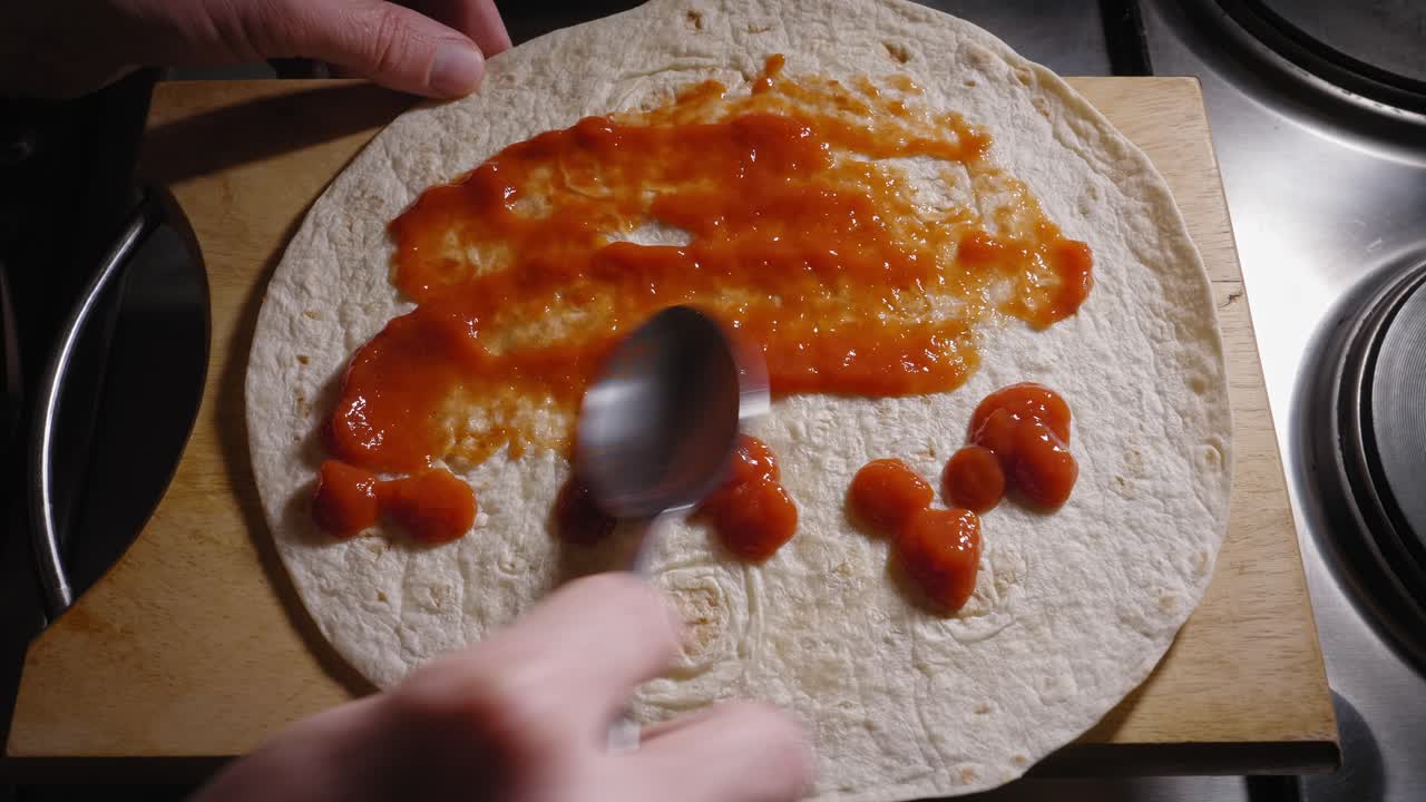 Una ripresa dall'alto delle mani di uno chef che spalma uniformemente una gustosa salsa di pomodoro su una tortilla con il dorso di un cucchiaio, la salsa che fornisce uno strato di sapore a una deliziosa tortilla Hot Dog.