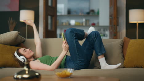 Relaxed girl laying couch listening music at home. Headphones lady enjoying song