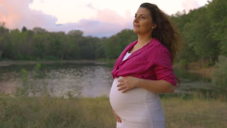 portrait of pregnant woman expectant mother walking alone in forest next to lake, attractive caucasian casual dressed mom stroking caress touching her belly with hands smiling, childbearing tenderness