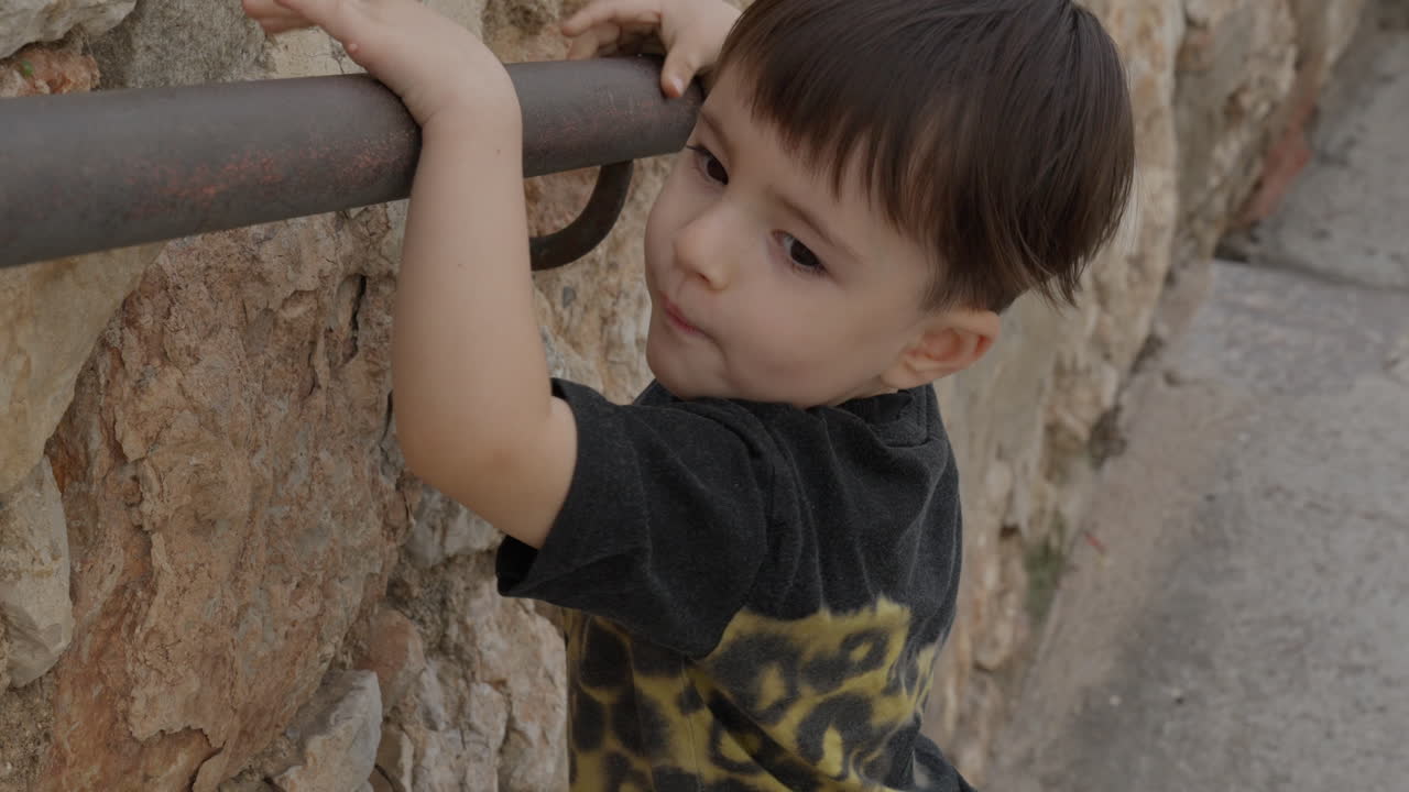 niño de pelo oscuro se aferra a la barandilla de metal mientras sube las escaleras de forma independiente y cuidadosamente