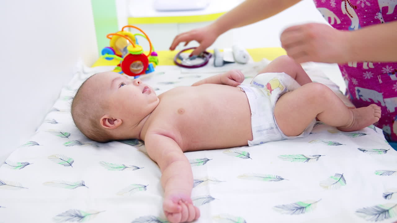 Cute naked baby in diaper lying on the doctor's table. Pediatrician standing in front of baby and kid watching the doctor curiously. Side view.