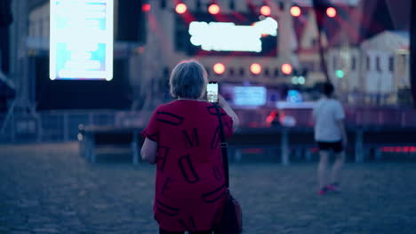 older caucasian woman making video with her smartphone of a stage with flickering red and white lights during a blue hour at night in an old historical town bardejov in slovakia in slow motion