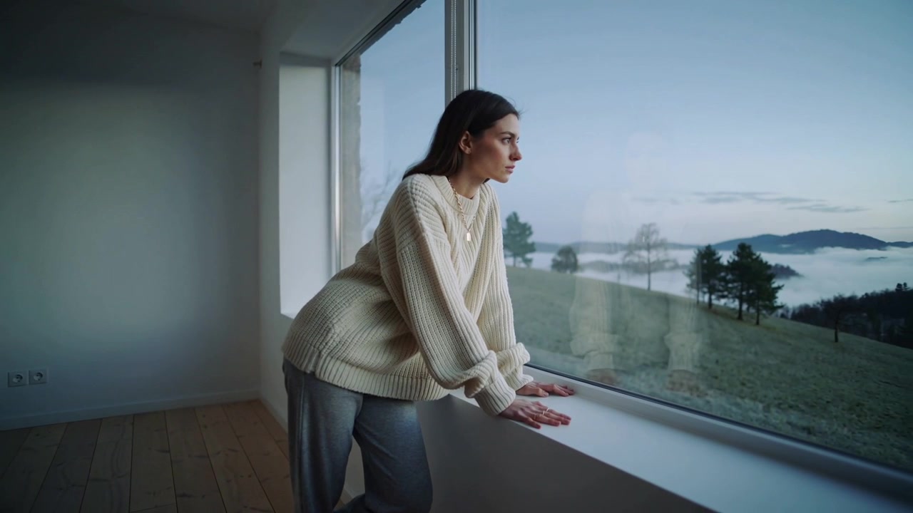 Una donna appoggiata, che indossa un maglione color crema, preme le mani sul davanzale della finestra di casa, mentre la nebbia attira lo sguardo all'esterno.