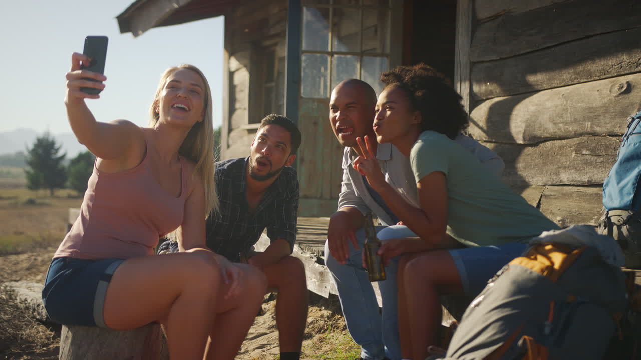 grupo de amigos de vacaciones sentados en el porche de una cabaña de campo bebiendo cerveza y tomando selfies
