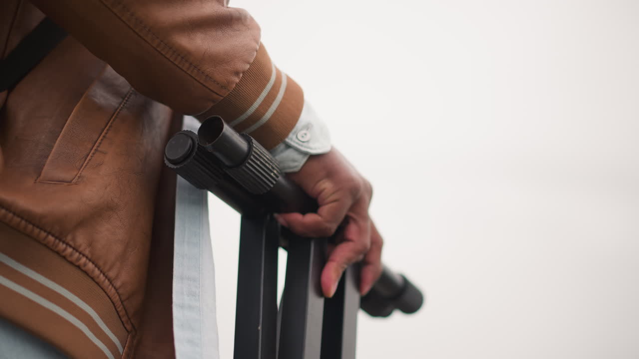 Closeup hand holding collapsed tripod against pale overcast sky, backpack and denim visible, leather jacket cuff, quiet preparation for street performance, intimate moody detail focusing on grip