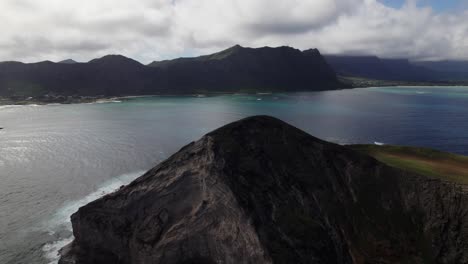 vista aérea girando alrededor de una pequeña isla montañosa frente a la costa de oahu hawai comenzando en el lado sombreado y girando para mostrar el lado iluminado por el sol de la formación volcánica