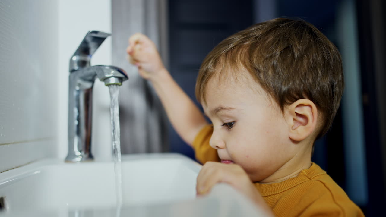 Cute little child opens the tap. Curious kid touches the flowing water and closes the faucet. Close up.