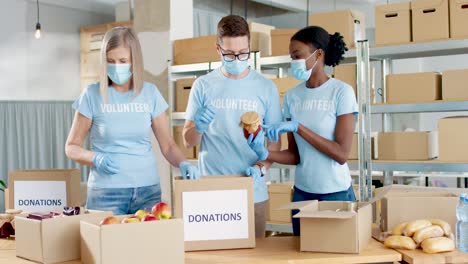 Grupo-Multiétnico-De-Voluntarios-En-Cajas-De-Embalaje-De-Máscaras-Faciales-Con-Comida-En-Almacén-De-Caridad