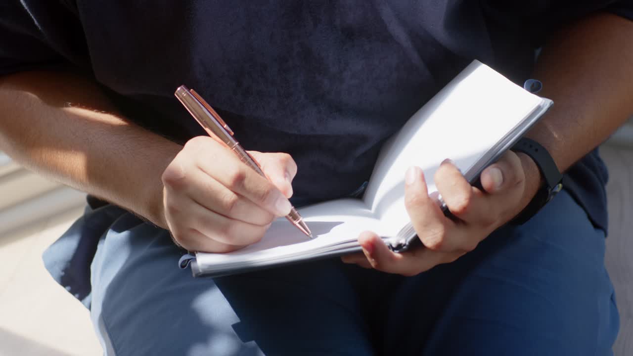 Mid-adult African American man positioning spiral notebook writing notes with rose-gold pen at home
