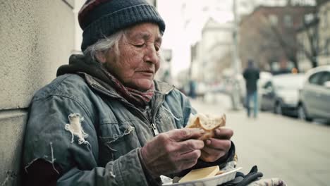 an elderly woman sits against a wall on a chilly city street, savoring a simple meal. she wears warm clothing, showing signs of wear, and highlights the daily struggles of the homeless.