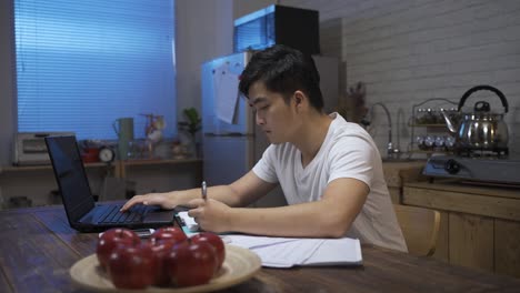 asian male working late from home in casual wear is writing note while doing accounting on the computer in the dining room at home.