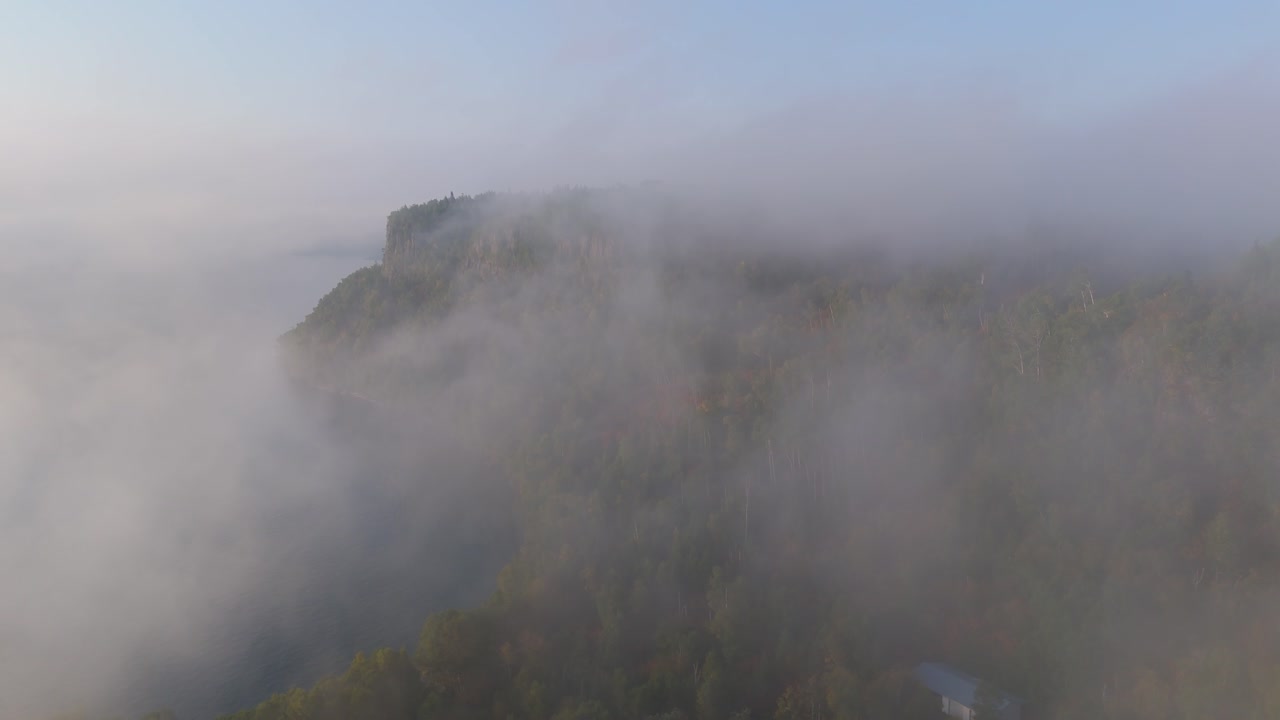 Dense morning fog drifts over the rugged shoreline of Lake Superior, revealing dramatic vertical cliffs and early autumn forest hues in Ontario, Canada. Aerial view