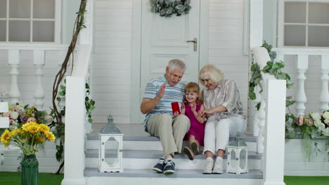 Senior-ouple-sitting-with-granddaughter-in-porch-at-home.-Using-mobile-phone-for-online-video-call