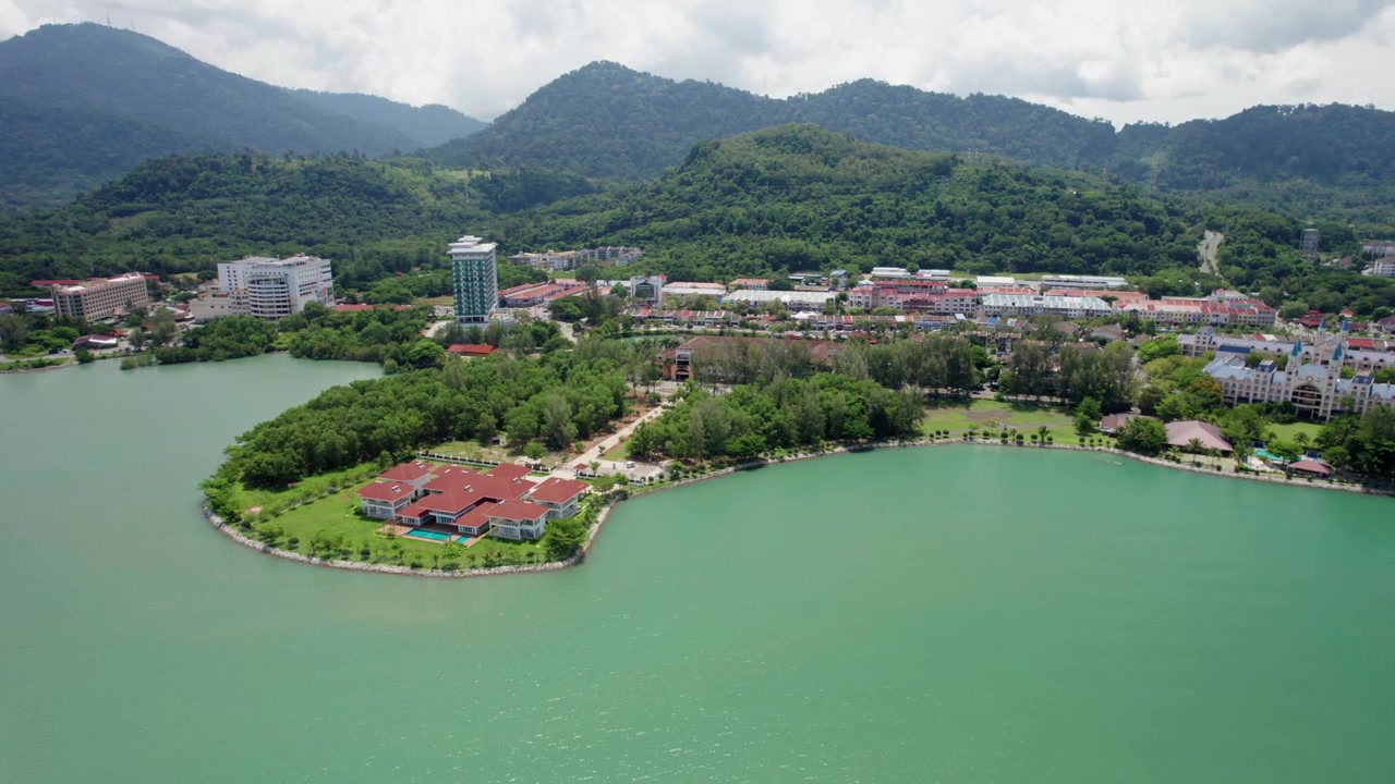 Malezya, Langkawi'nin eşsiz coğrafyasını vurgulayan, yağmur ormanlarıyla kaplı dağlar ve sahil binalarıyla çevrili huzurlu bir tropikal koyu gösteren drone manzarası.