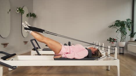 woman in workout clothes performs leg exercises on pilates reformer at gym. wide static shot