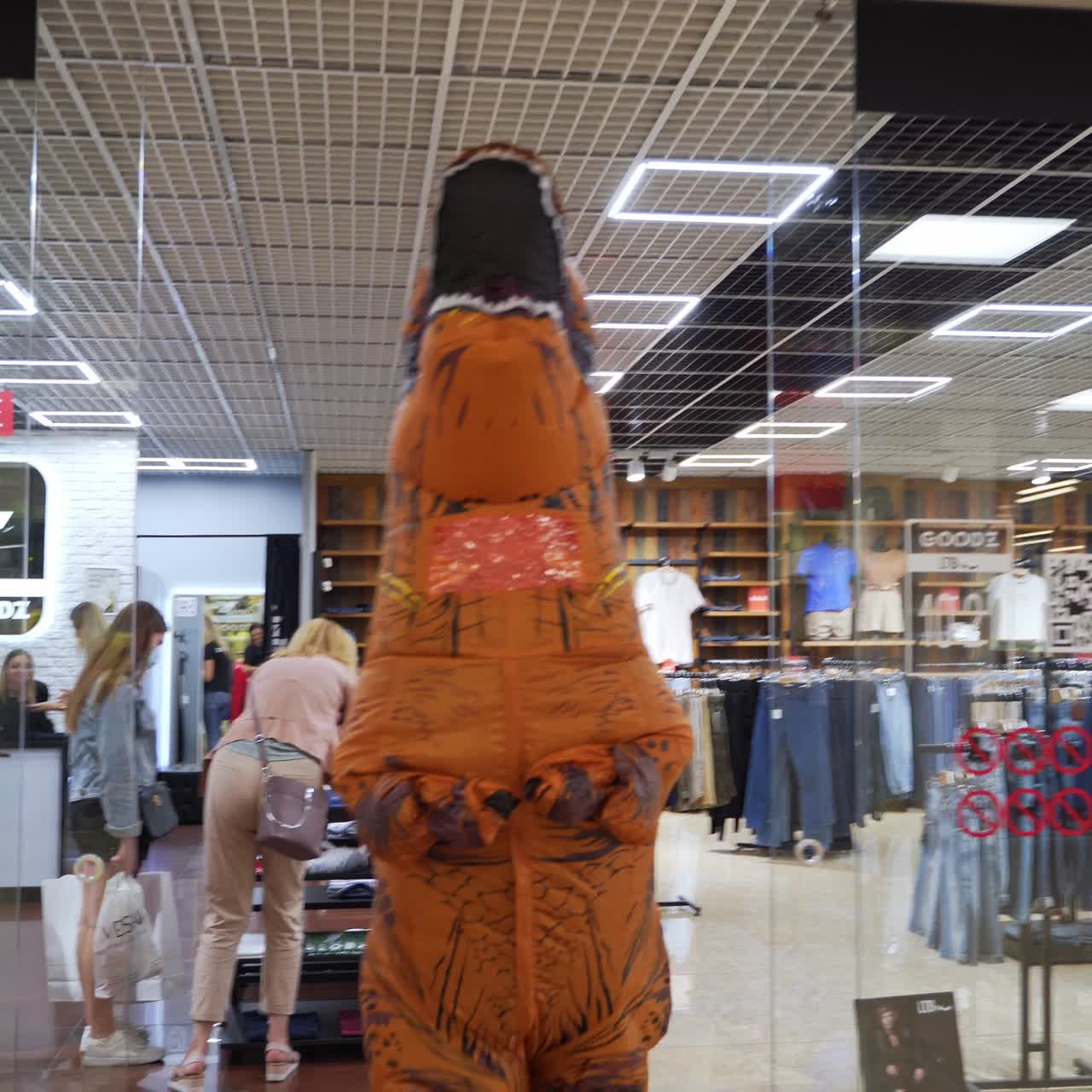 Man in orange funny spotty costume of dinosaur leaves the clothes shop. T-Rex runs quickly by the spacious hall of modern shopping mall