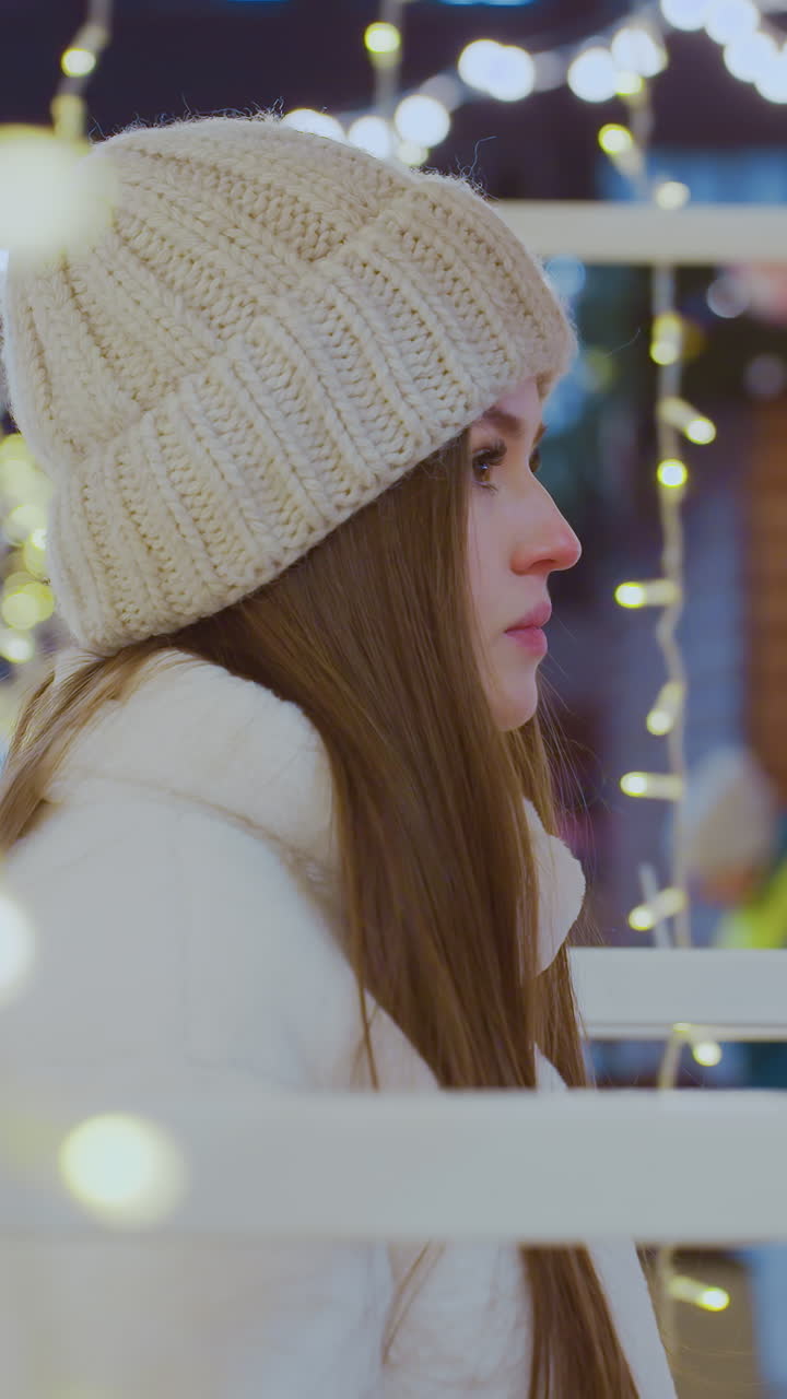 Elegant woman in white beanie and winter jacket walking past glowing golden holiday lights at night, festive atmosphere with decorative string lights illuminating surroundings