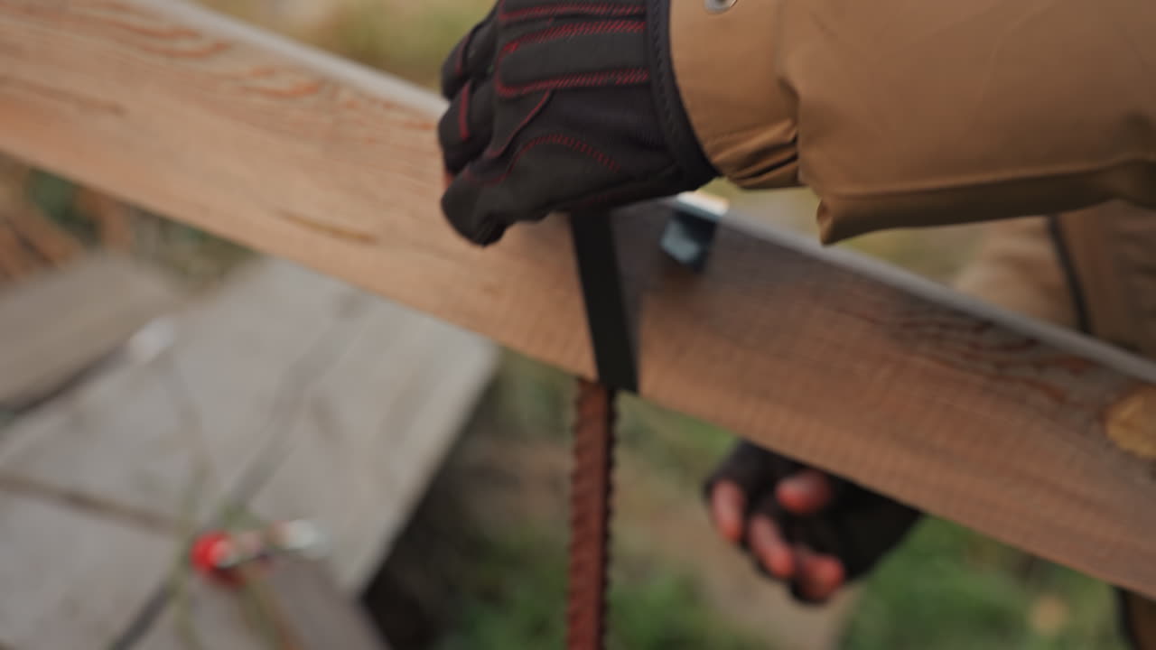 Behandschuhte Hände reparieren einen Holzbalken im Freien. Ein sorgfältiger Techniker befestigt und sichert einen Gurt an einer Holzplanke. Nahaufnahme von Schutzhandschuhen und Jacke, methodische Wartungsprüfung, konzentrierte Anspannung.