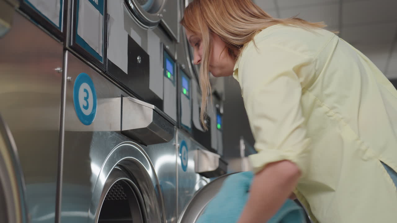 Business woman carries basket of clothes toward industrial washer, door open, preparing to load garments into drum inside laundromat, stainless panels visible, hygiene routine, clean interior scene