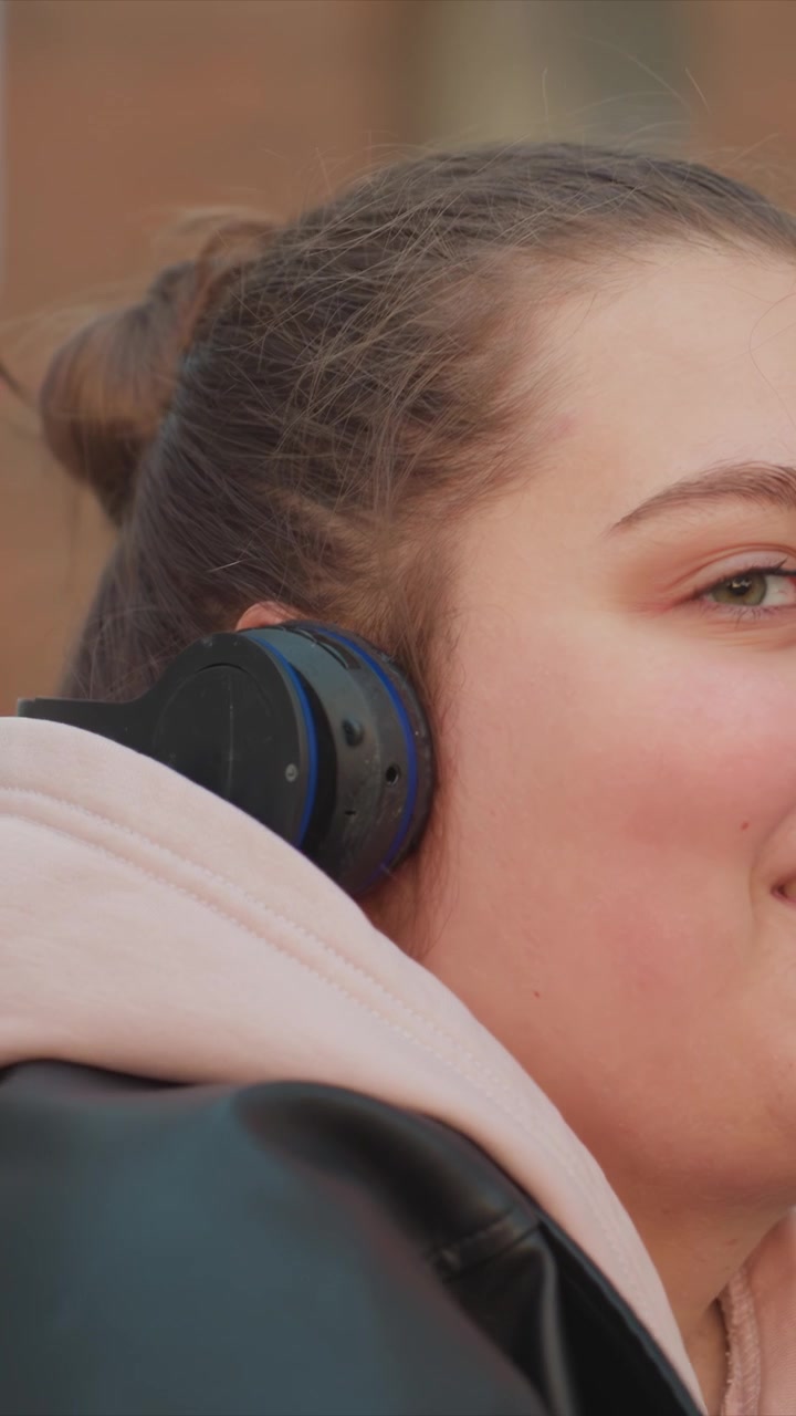 Ragazza con capelli scuri raccolti in uno chignon che indossa una giacca di pelle nera. Manipola una fascia verso l'orecchio e sposta la fotocamera. Un primo piano mostra un movimento minimo della testa sullo sfondo di una città all'aperto.