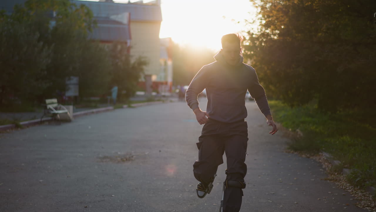 한 남자가 해가 지는 동안 주거 거리를 따라 기둥 위에서 조하고 있으며, 중간 거리에서 포착되었습니다. 회색 갈색 스웨트 셔츠와 어두운 화물 바지를 입고, 심각한 표정과 반이는 황금색 빛으로
