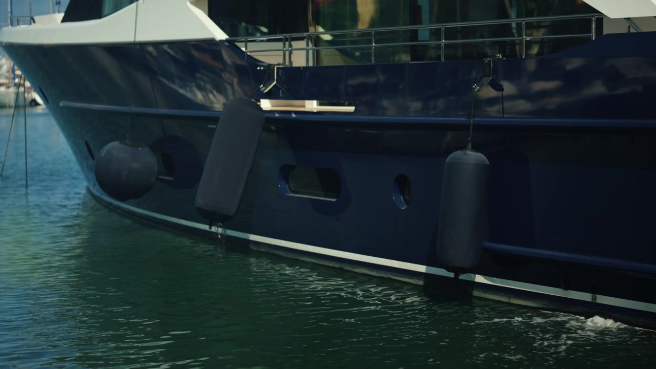 Blauer Rumpf einer modernen Luxusyacht mit mehreren hängenden Bootsfendern, in einem ruhigen Hafen festgemacht. Repräsentiert Reichtum und Luxusreisen.