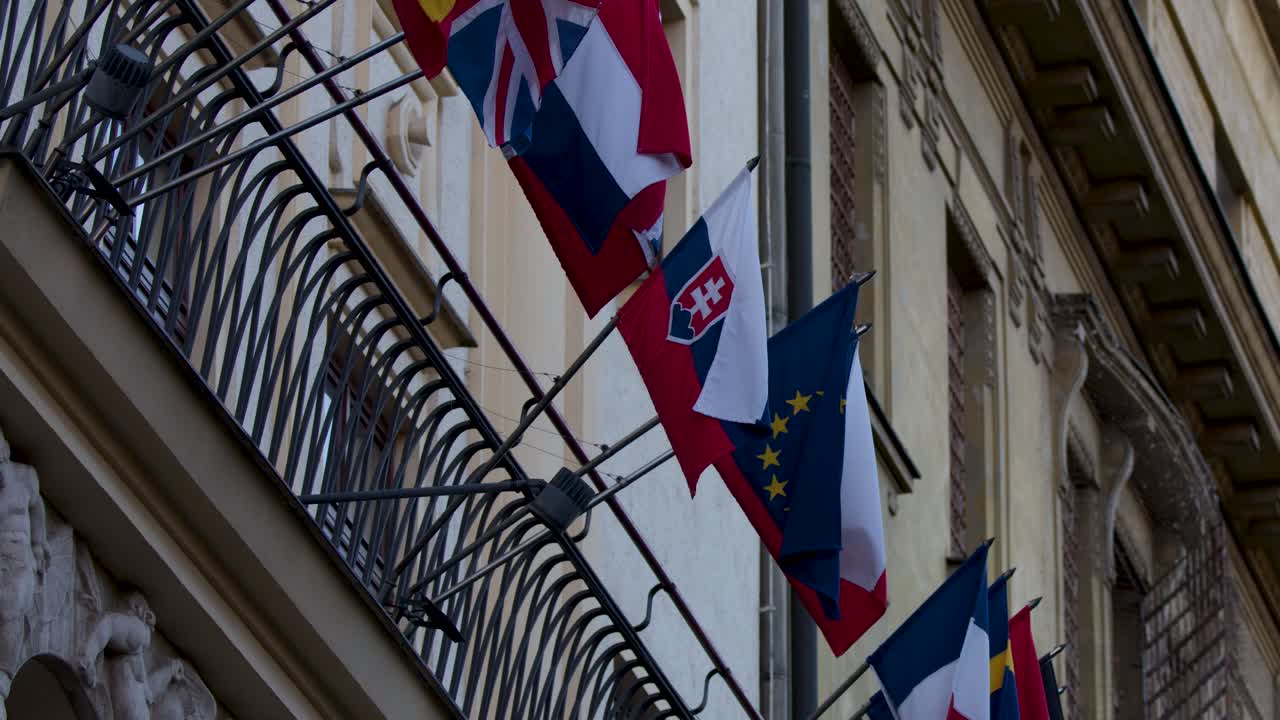 Multiple international flags flutter on ornate balcony, overcast daylight, slight upward camera pan, city center