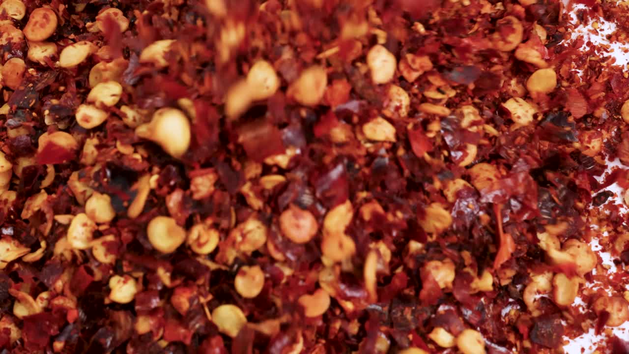 Dried chili flakes cascade onto a white surface, creating a vibrant, textured pattern in bright lighting