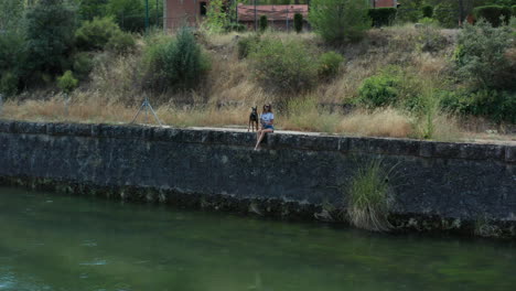 Mujer-Joven-Sentada-Con-Su-Perro-A-Lo-Largo-De-La-Orilla-Del-Río