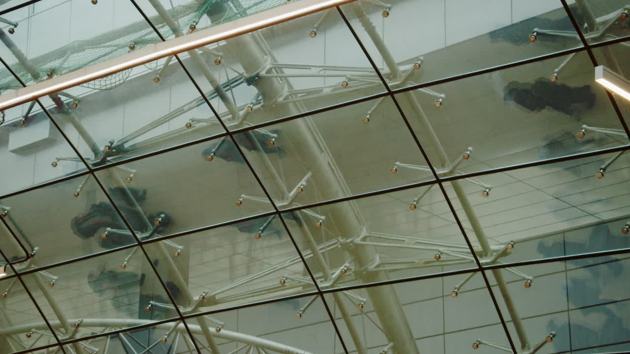 Abstract view of a modern glass roof with reflections of people walking above. Architectural structure with steel beams and transparent panels, creating an urban, minimal and contemporary atmosphere