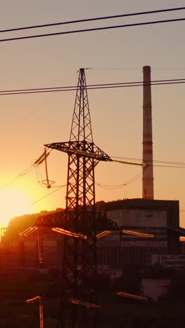 electricity pylons at sunset. high-voltage electric tower on industrial pipe background. transmission lines near the factory with dirty smoke into the air in the evening. vertical video
