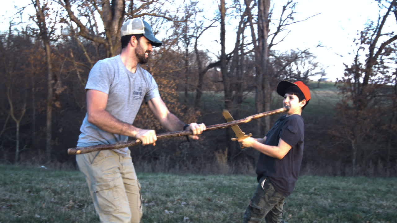 Father and son engage in playful sword fight with sticks in the woods at dusk, showing family bonding