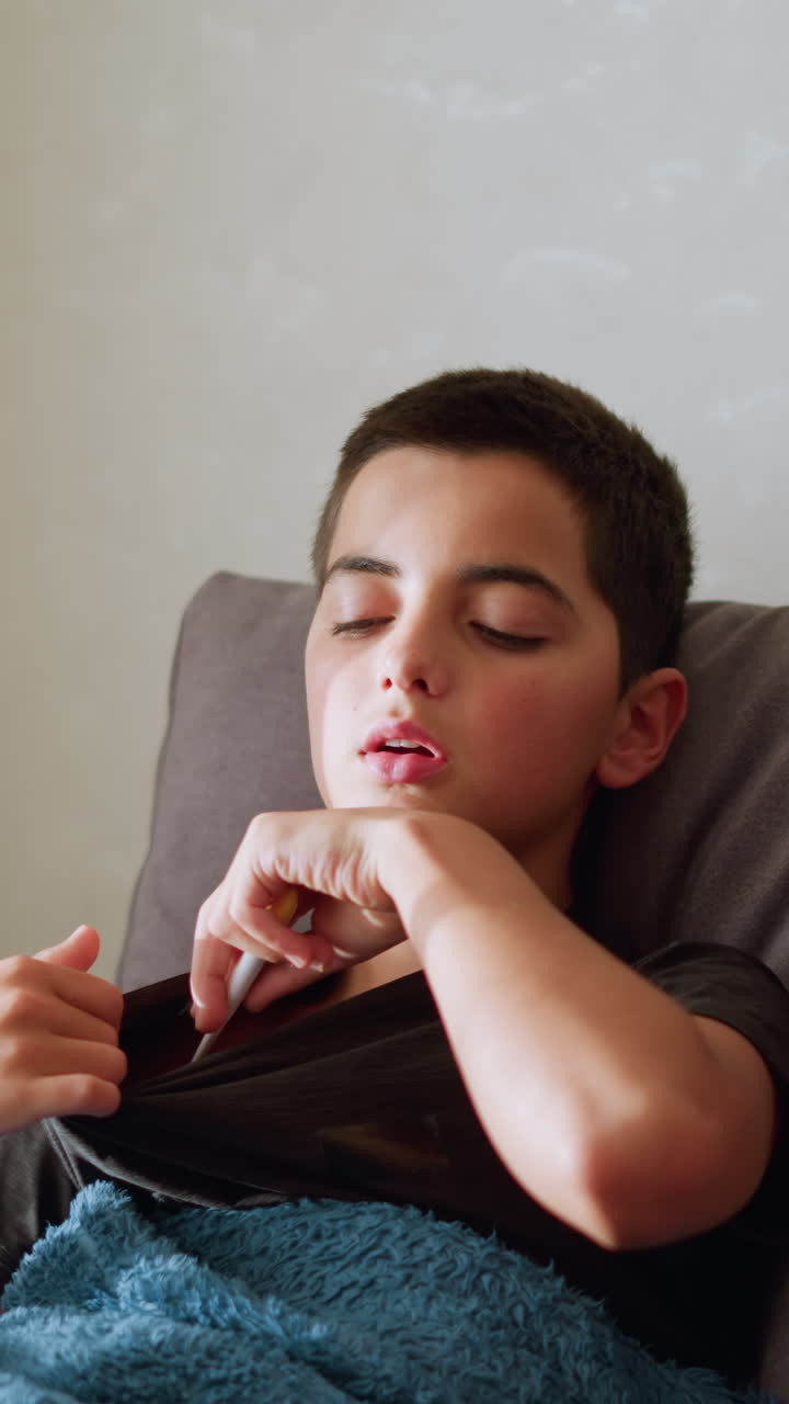 Girl with rope around her neck hands thermometer to sick brother sitting on couch to check his temperature as he places it under his armpit, soft light filters through window