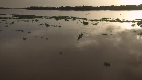 Vista-Aérea-De-Una-Pequeña-Canoa-Indígena-Cruzando-Un-Montículo-De-Algas-Flotantes-En-El-Río-Orinoco-Durante-La-Puesta-De-Sol