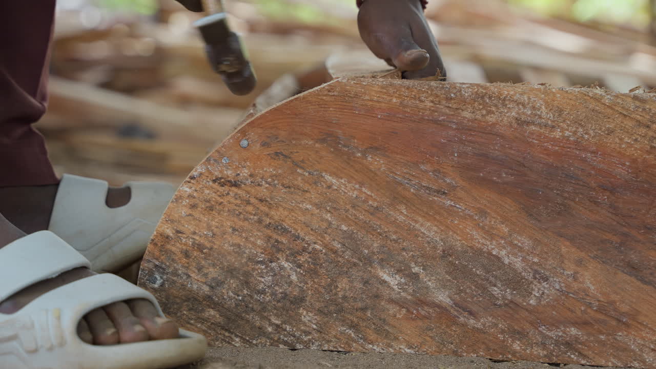 Mani scure rifiniscono legno texturizzato. Scena all'aperto che mostra una falegnameria dettagliata con sandali bianchi sul terreno. Artigiano paziente usa uno strumento manuale per rifinire e lucidare una superficie di legno texturizzata all'aperto.