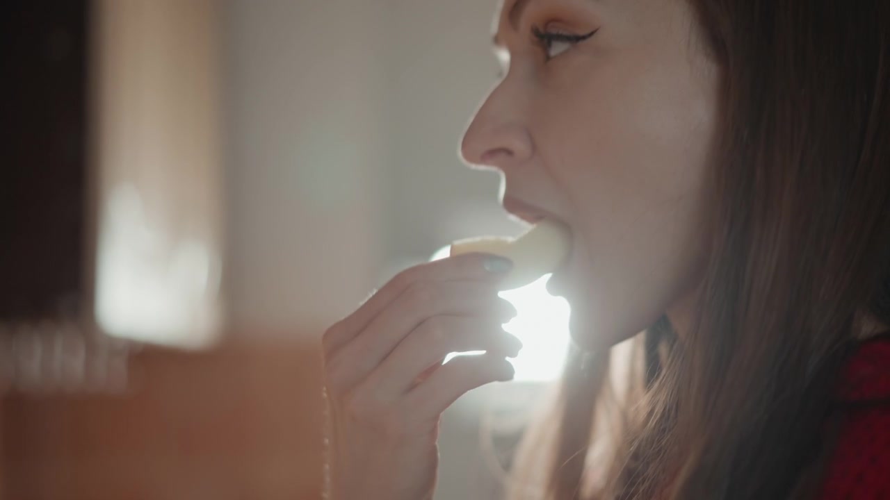 Vista di profilo di una donna che indossa un maglione rosso, con gli occhi chiusi durante una delicata espirazione accanto a una luminosa finestra della cucina; morbida retroilluminazione, rimlight e un leggero bagliore enfatizzano la calma pacifica del mattino.