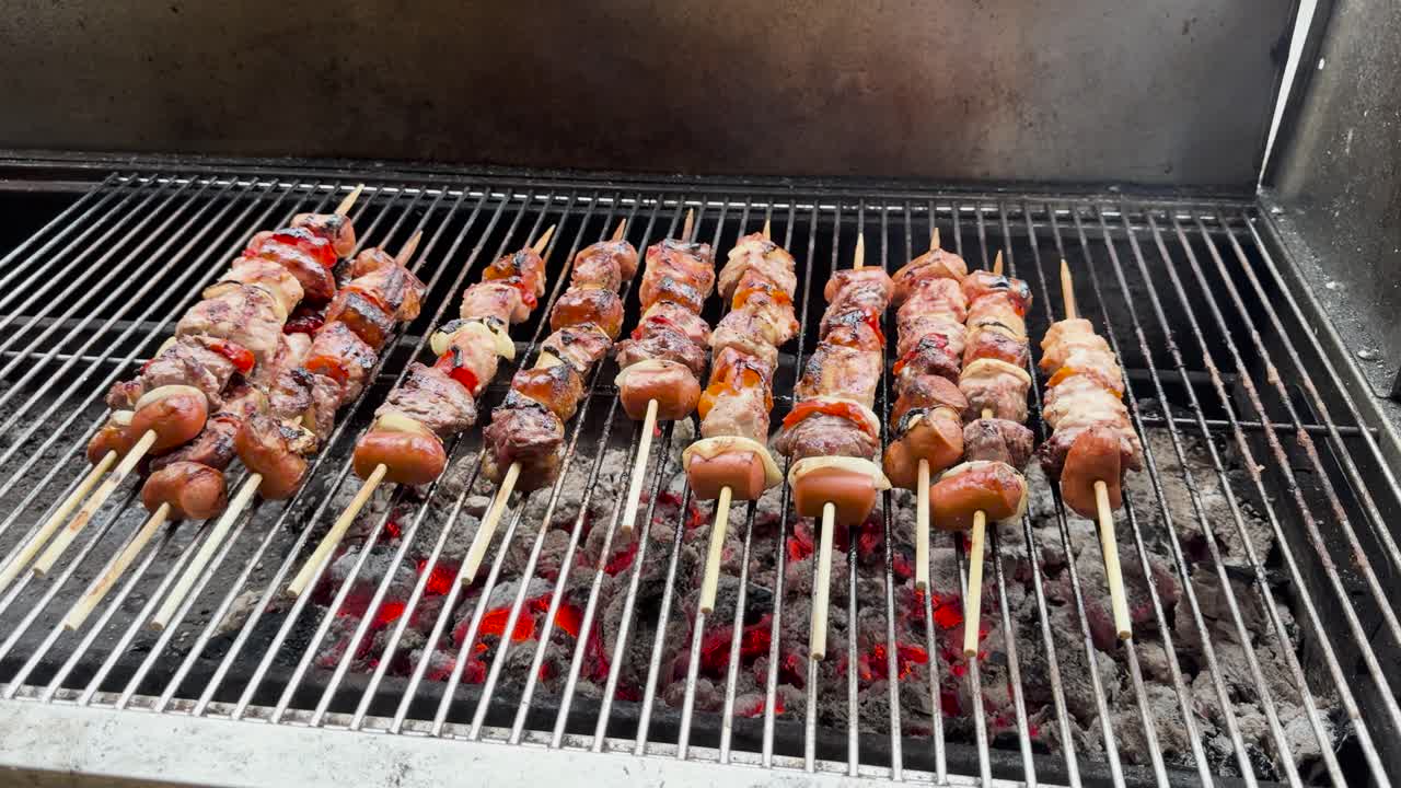Static handheld video of meat skewers on a charcoal grill, with red peppers, white onions, and sausages. Glowing red coals and light smoke create a rustic, savory scene.