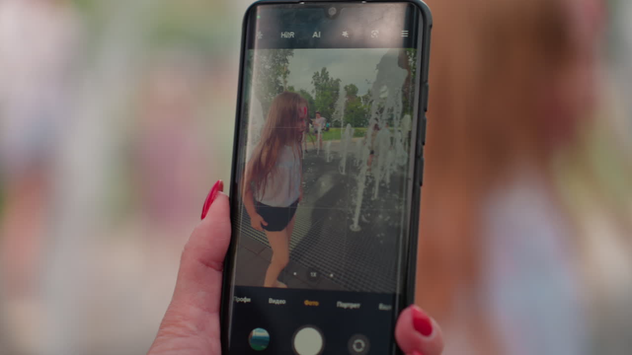 close up female hand holding phone camera capturing girl playing with water fountain, bare legs running through jets, joyful summer park scene, splash motion and candid family fun