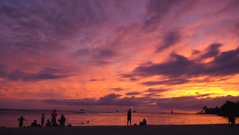 Menschen-Stehen-Am-Strand-Und-Beobachten-Die-Farben-Eines-Tiefroten-Sonnenuntergangs,-Die-Sich-Im-Ozean-Spiegeln