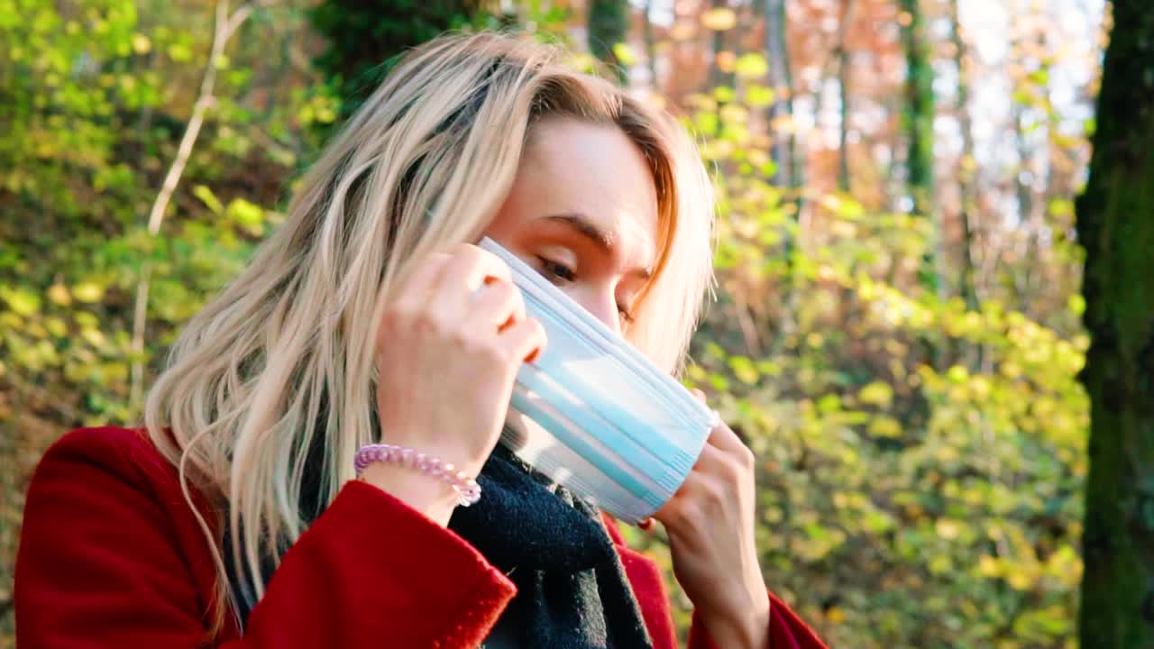 SLOWMOTION Ritratto di una giovane e bellissima donna che indossa una mascherina di sicurezza anti-Corona in un bosco autunnale dai colori arancioni e marroni, mentre porta un cappotto rosso