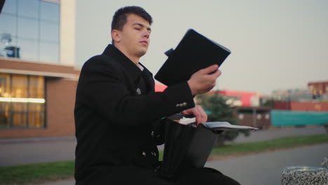 man seated outdoors going through briefcase searching for something, taking out tablet from bag, with background featuring modern urban building and city street environment