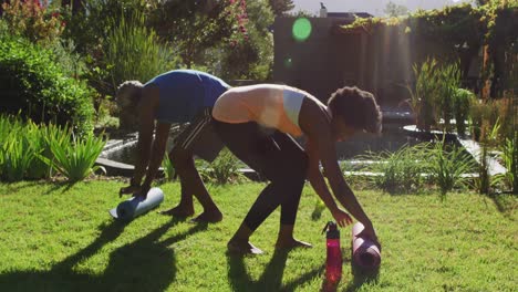 pareja de ancianos afroamericanos haciendo ejercicio al aire libre enrollando esteras de yoga en un jardín soleado