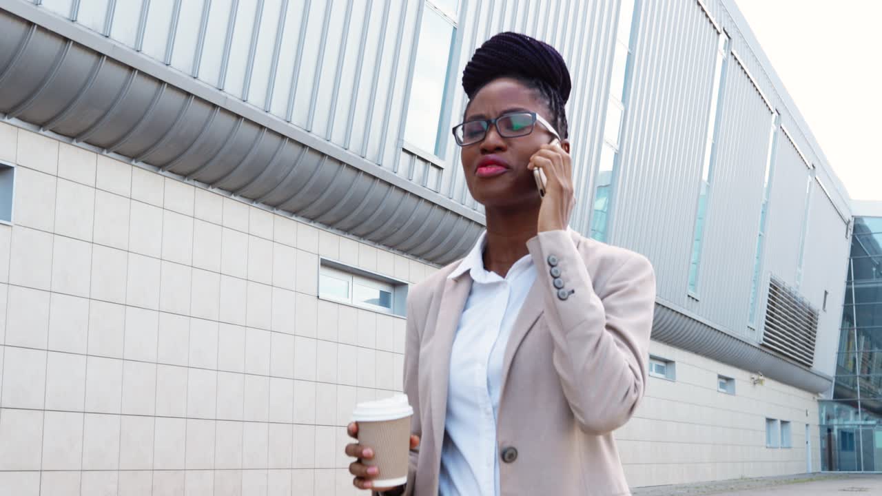 mujer de negocios afroamericana con ropa y gafas elegantes hablando por teléfono inteligente y tomando café para ir a la calle