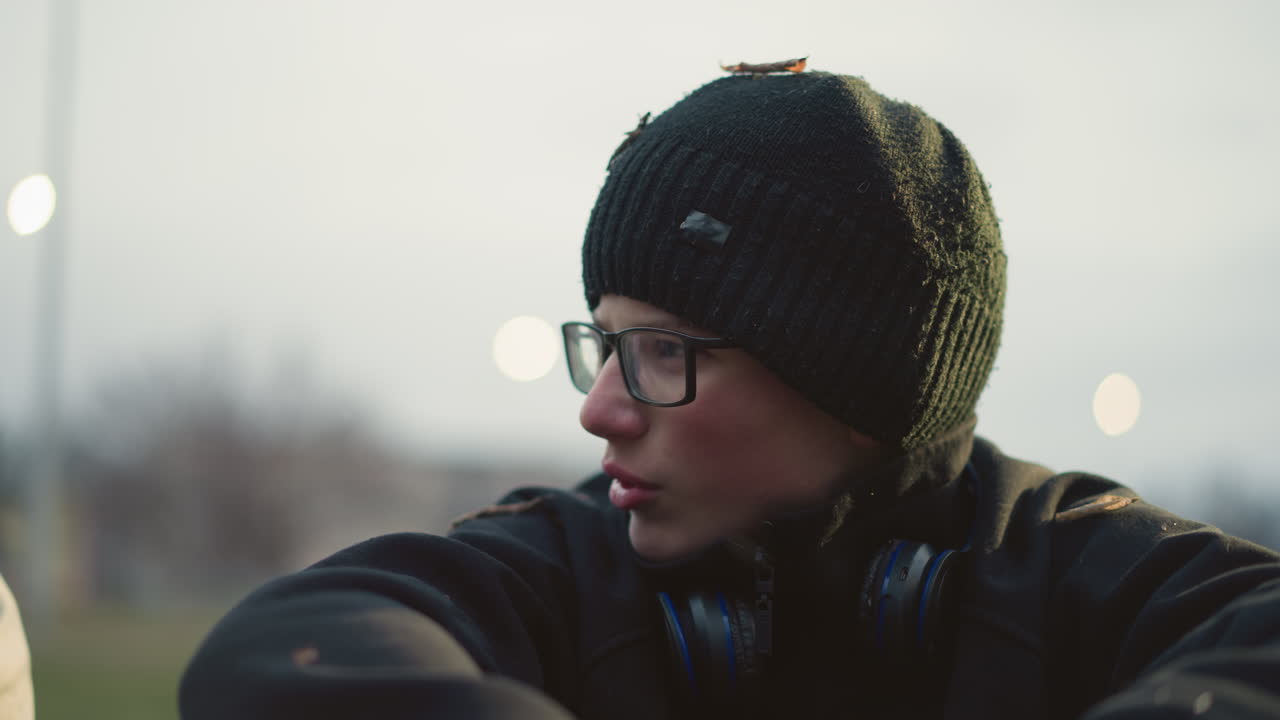 Close-up of a young boy wearing glasses and a black beanie with grass particles on his head and headset on his neck, engaged in a discussion with someone