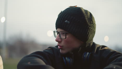 close-up of a young boy wearing glasses and a black beanie with grass particles on his head and headset on his neck, engaged in a discussion with someone